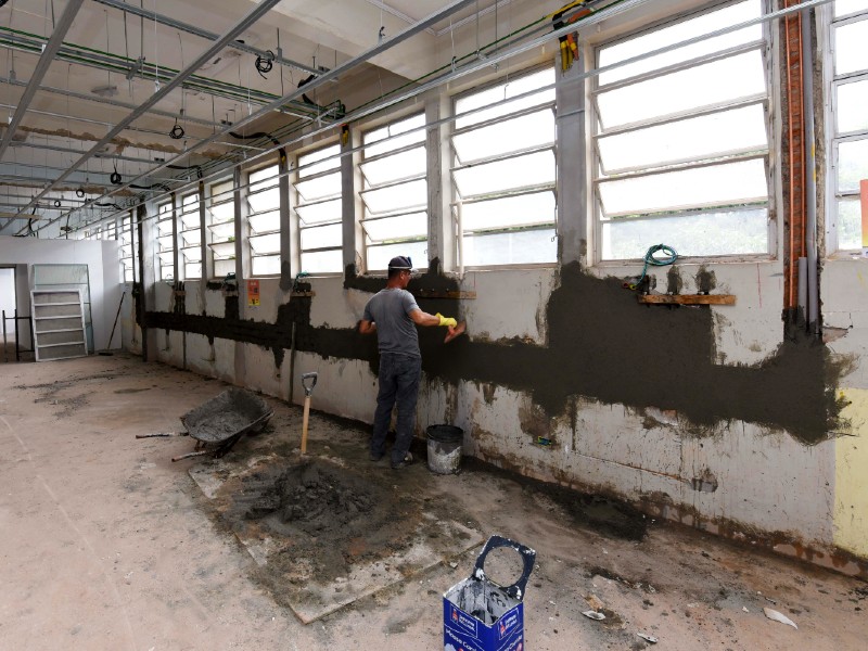Foto da obra da UPA Vergueiro. Um homem aplica uma massa de cimento na parede. Há ferramentas no chão e estruturas para iluminação que saem do teto